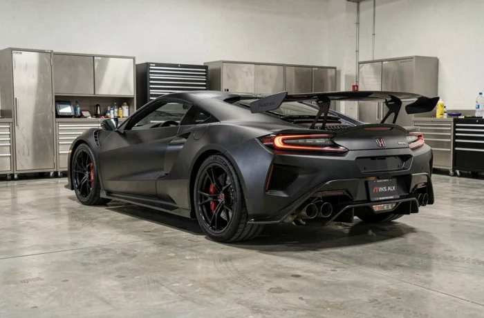 Honda NSX deuxième génération noire dans un garage motorsport moderne, vue trois quarts arrière soulignant l'aérodynamique agressive et les sorties d'échappement
